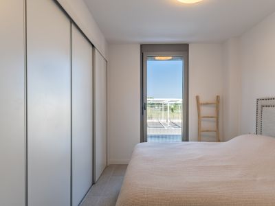Helles Schlafzimmer mit weißem Einbauschrank und Schiebetüren, Fliesenboden und direktem Zugang zum Außenbereich durch eine Glastür. Bett mit gepolstertem Kopfteil.