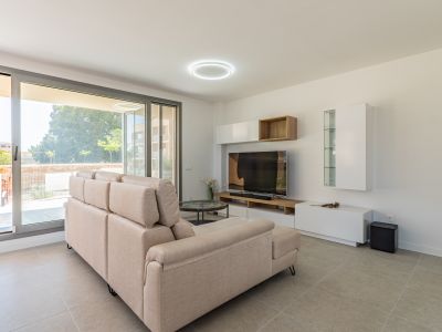 Helles Wohnzimmer mit beigem Ecksofa, hellgrauem Fliesenboden und großen Glasschiebetüren zum Außenbereich. Moderne TV-Möbelkombination mit weißen und Holzregalen.