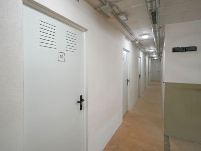 Langer Lagerflur mit weißen Türen, Lüftungsschlitzen und schwarzen Griffen. Die Decke zeigt sichtbare Lüftungskanäle und Einbauleuchten über einem Betonboden.
