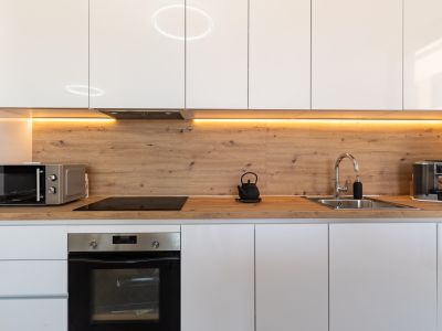Moderne Küche mit glänzenden weißen Schränken, Arbeitsplatte und Rückwand in Holzoptik sowie warmer LED-Unterbauleuchte. Ausgestattet mit Backofen, Induktionskochfeld und Mikrowelle.