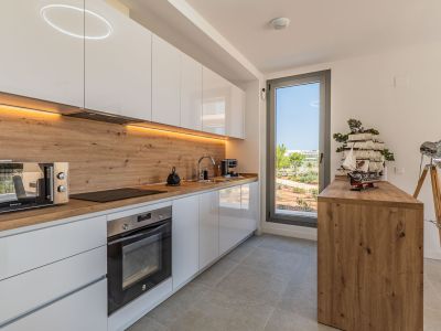 Moderne Küche mit hochglänzenden weißen Schränken, Arbeitsplatte und Spritzschutz in Holzoptik. Ausgestattet mit integrierten Geräten, LED-Beleuchtung und Fenster mit Blick ins Grüne.