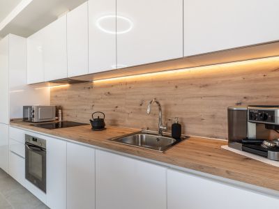 Moderne Küche mit hochglänzenden weißen Schränken, heller Holzoptik-Arbeitsplatte und passender Rückwand. Ausgestattet mit Einbaubackofen, Induktionskochfeld und Edelstahlspüle, beleuchtet durch LED-Unterbauleuchten.