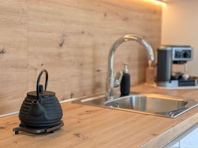Moderne Küchenarbeitsplatte und Rückwand in heller Holzoptik. Edelstahlspüle mit Chromhahn, beleuchtet durch LED-Unterschrankleuchten. Eine Teekanne aus Gusseisen steht im Vordergrund.