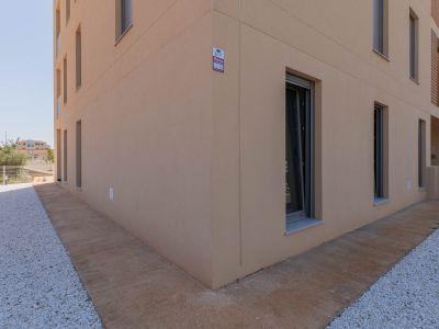 Moderner Gebäudeaußenbereich mit heller beigefarbener Putzfassade und dunkel gerahmten Fenstern. Der Boden zeigt einen strukturierten Betonweg neben einer weißen Kiesfläche, mit sichtbarer Alarmanlage.