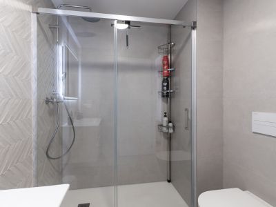 Modernes Badezimmer mit Glasduschkabine und weißer Duschwanne. Die Duschwand ist mit hellgrauen Fliesen im Fischgrätmuster versehen. Regendusche und wandhängendes WC sind sichtbar.