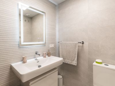 Modernes Badezimmer mit rechteckigem weißen Waschbecken, Wandarmatur, beleuchtetem Spiegel und hellgrauen strukturierten Wandfliesen.