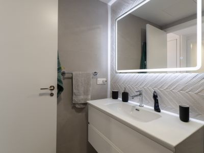 Modernes Badezimmer mit schwebendem Waschtisch, Chromarmatur und beleuchtetem Spiegel. Auffällig sind die strukturierten Wandfliesen im Fischgrätmuster.