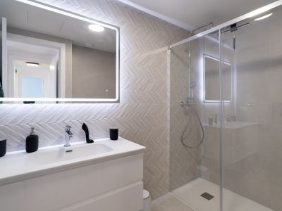 Modernes Badezimmer mit weißem Waschtisch, beleuchtetem Spiegel und Wandfliesen im Fischgrätmuster. Die Glasdusche verfügt über eine Regendusche und Chromarmaturen.