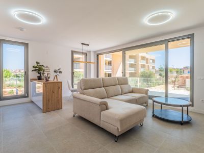 Offener Wohn- und Essbereich mit beigem Ecksofa und ovalem Couchtisch. Große Glasfronten sorgen für viel Tageslicht und Blick auf die Wohnanlage. Offene Küche mit Holztheke.