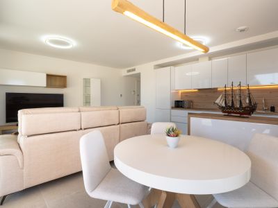 Offener Wohn- und Essbereich mit integrierter Küche. Helles Sofa, weißer ovaler Esstisch und moderne Küche mit hochglänzenden weißen Schränken, Holzdetails und indirekter Beleuchtung.