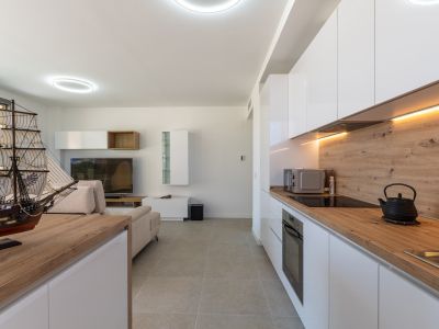 Offener Wohn- und Essbereich mit moderner Küche. Weiße Hochglanzschränke, helle Arbeitsplatten in Holzoptik und integrierte Geräte werden durch warmes LED-Licht und graue Fliesenböden betont.