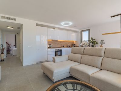 Offener Wohn- und Küchenbereich mit beigem Ecksofa, modernen weißen Hochglanzschränken und heller Holzrückwand. Fliesenboden und Tageslicht durch große Fenster.