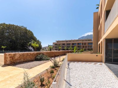 Privater Erdgeschossgarten mit weißem Kies und Trittsteinen, umzäunt von einem niedrigen weißen Zaun und einer Steinmauer. Blick auf moderne Gebäude und entfernte Berge.