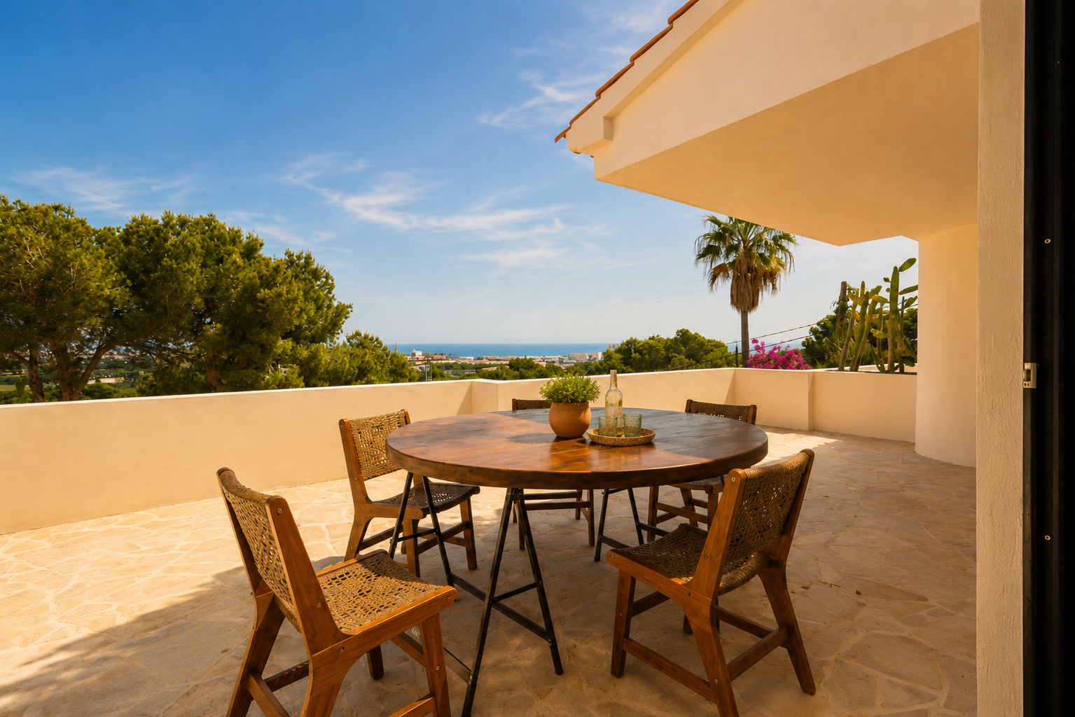 Außenterrasse mit rundem Holztisch und geflochtenen Stühlen auf Steinplatten, mit Panoramablick auf das Meer und die Küste.