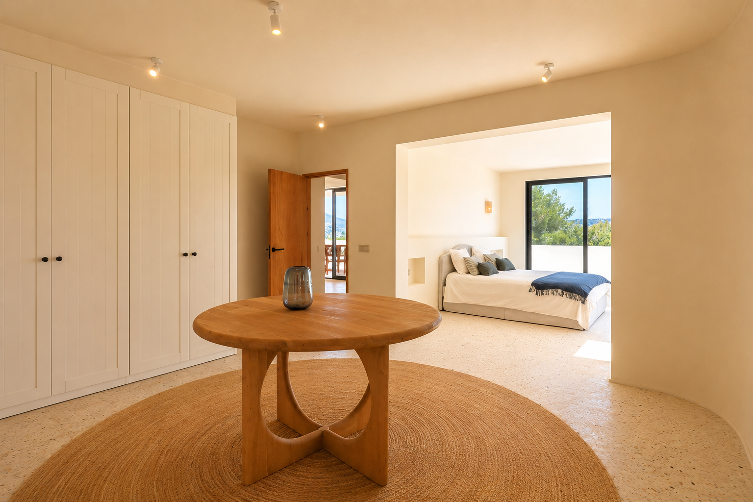 Dieser helle Übergangsbereich verfügt über einen runden Holztisch auf einem Juteteppich, flankiert von weißen Einbauschränken. Er führt zu einem Schlafzimmer mit grüner Aussicht.