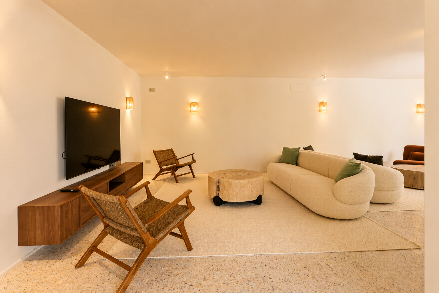 Ein moderner Wohnbereich mit geschwungenem cremefarbenem Sofa, rustikalem Holztisch und Korbsesseln auf Terrazzoboden, beleuchtet von warmen Wandleuchten.