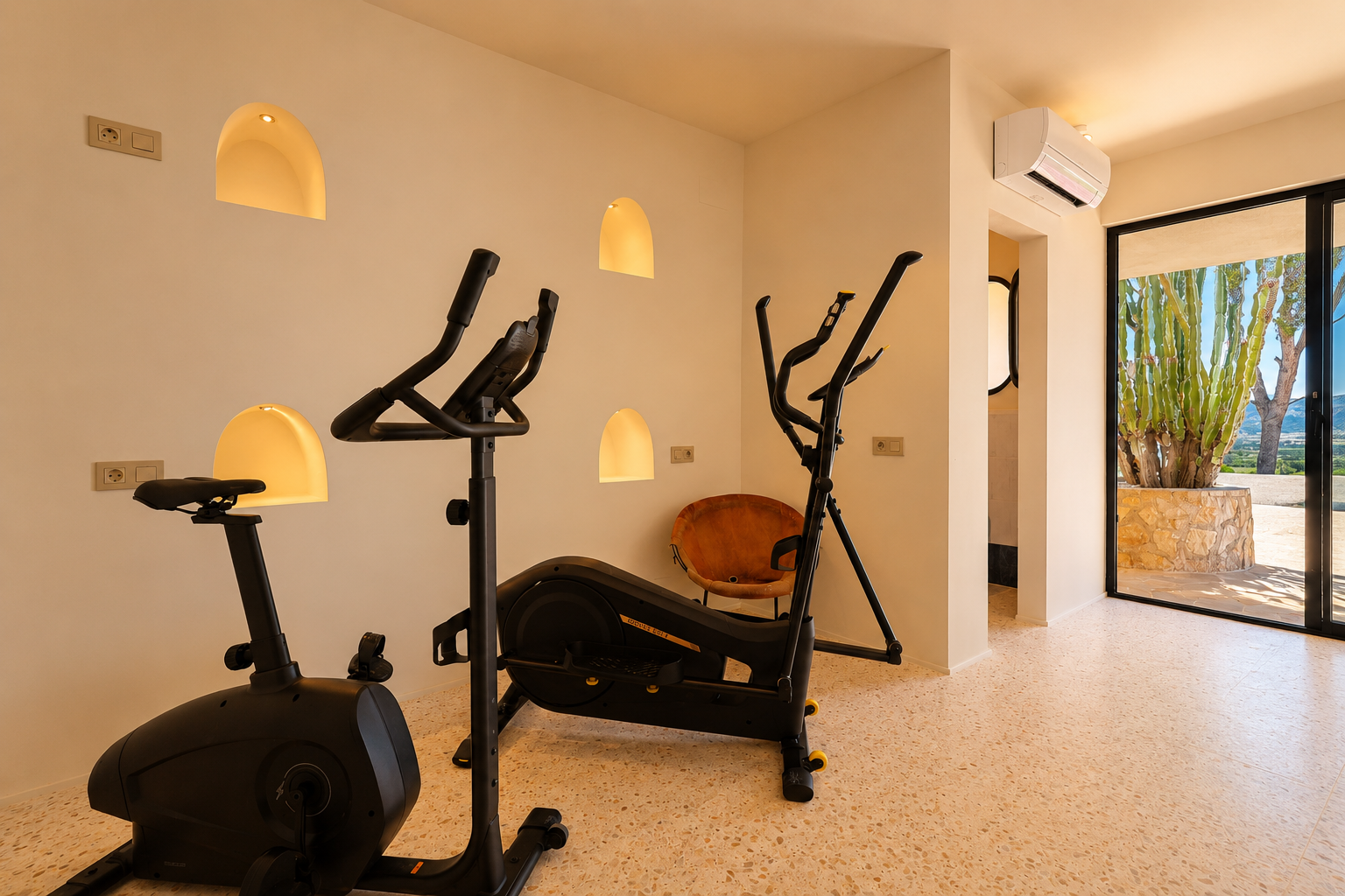 Fitnessbereich mit Heimtrainer und Crosstrainer, beleuchteten Bogennischen in der Wand und Blick auf einen Kaktusgarten und die Berge.