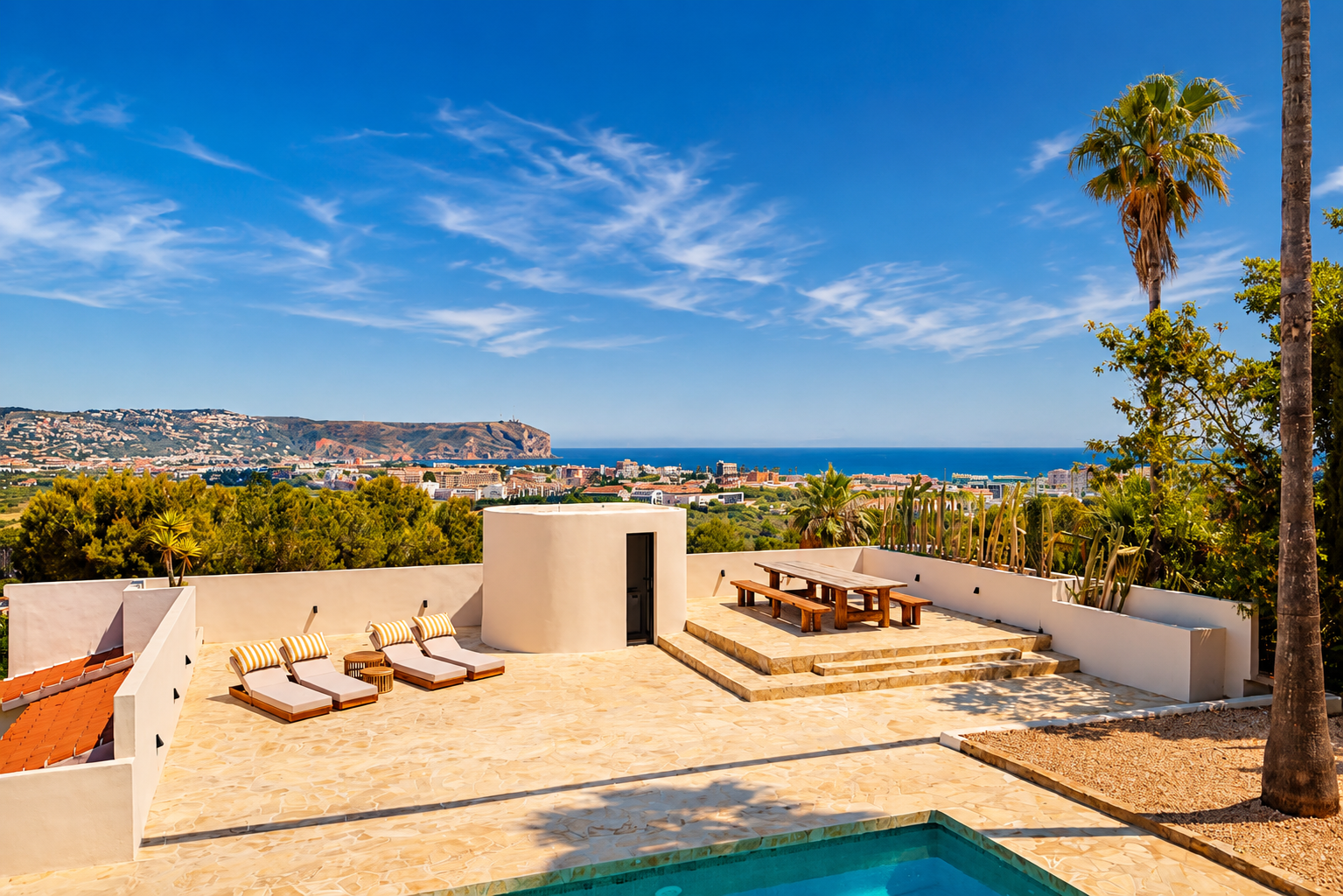 Großzügige Steinterrasse mit Pool, Sonnenliegen und erhöhtem Essbereich aus Holz, mit Panoramablick auf die Bucht von Jávea und die Küste unter blauem Himmel.