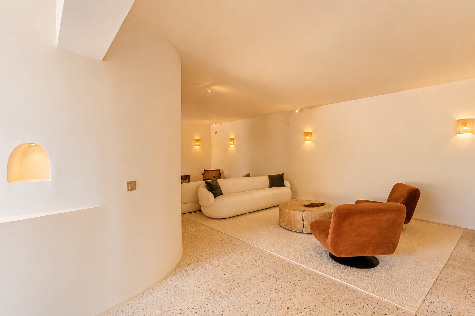 Moderner Loungebereich mit geschwungenem cremefarbenen Sofa, zwei rostfarbenen Sesseln und rustikalem Holztisch auf hellem Terrazzoboden. Geflochtene Wandleuchten sorgen für warmes Licht.