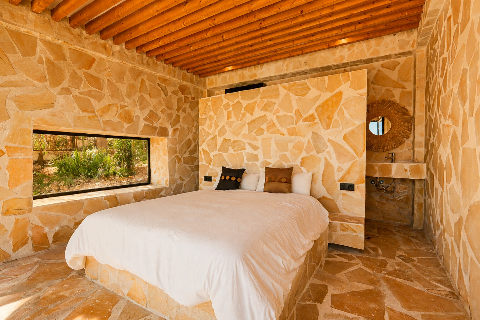 Schlafzimmer mit unregelmäßigen Natursteinwänden und -boden, Holzbalkendecke und einem horizontalen Fenster mit Gartenblick. Ein integriertes Steinwaschbecken mit Rattan-Spiegel ist sichtbar.