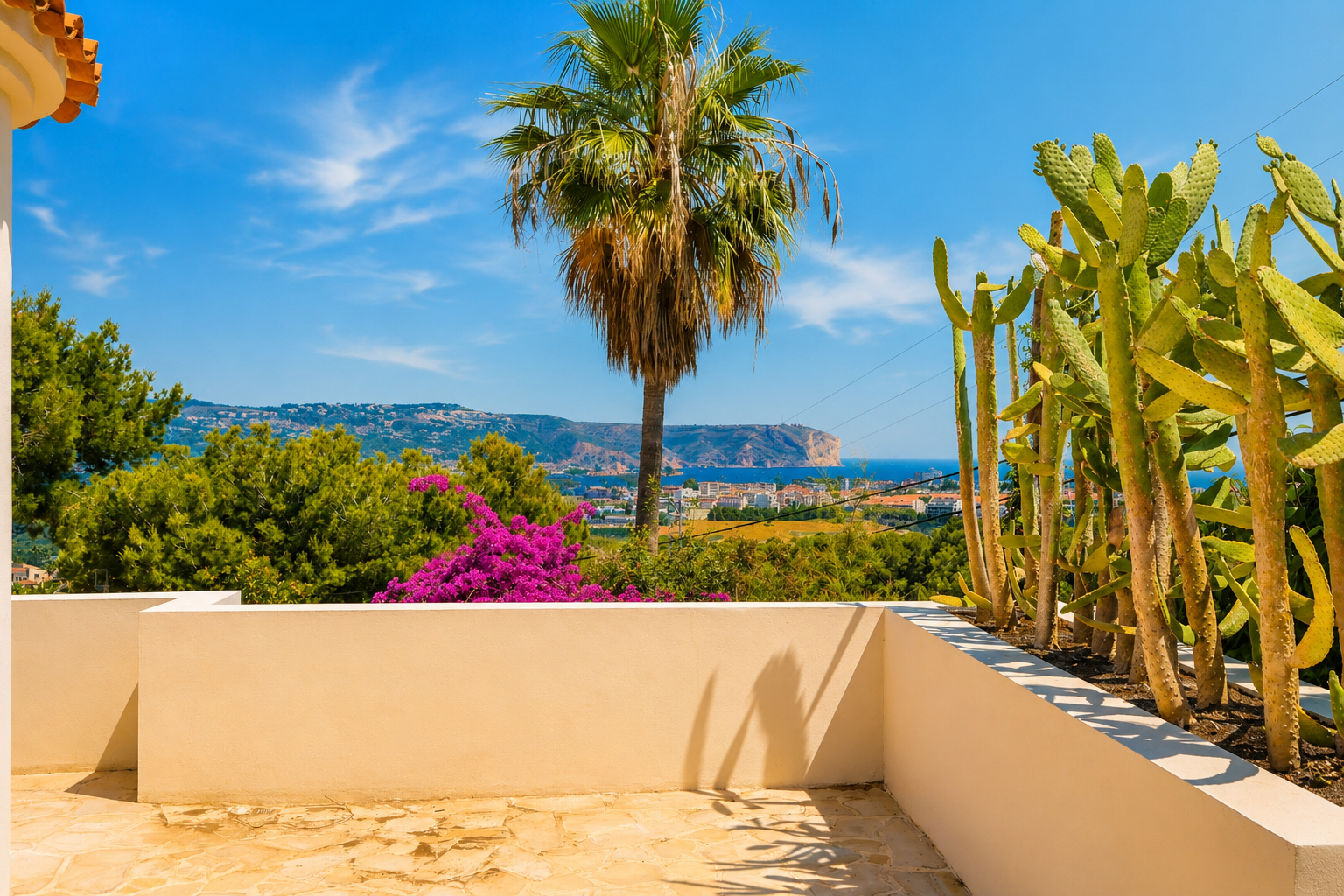 Terrasse mit unregelmäßigem Steinboden und niedriger beiger Mauer. Bietet einen Kaktusgarten und Panoramablicke auf das Mittelmeer, die Küste und ein entferntes Kap bei blauem Himmel.