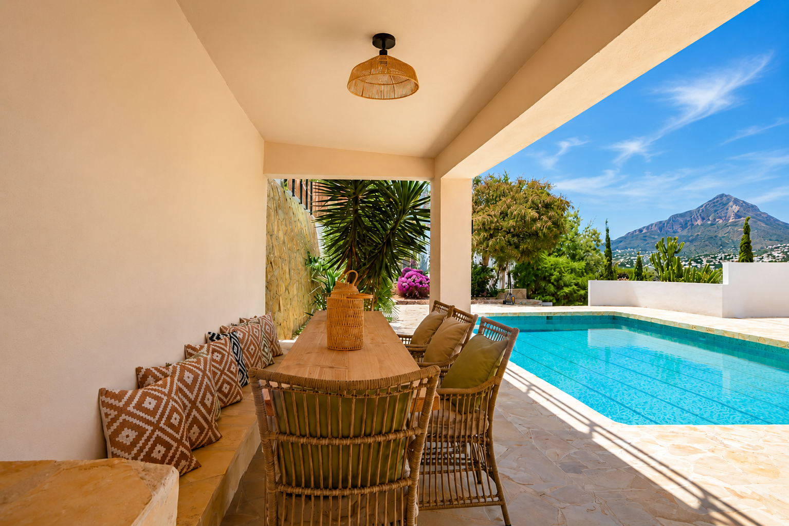 Überdachter Essbereich im Freien mit Holztisch und Korbstühlen, direkt am Pool gelegen. Panoramablick auf den Montgó und mediterrane Pflanzenwelt auf Steinboden.
