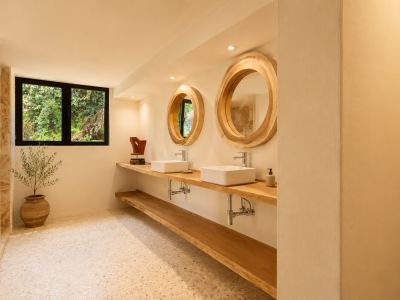 Badezimmer mit doppeltem Holzwaschtisch, zwei weißen rechteckigen Waschbecken und großen runden Spiegeln mit Naturholzrahmen. Ein Fenster blickt auf üppiges Grün, passend zum hellen Terrazzoboden.