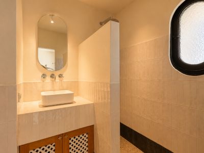 Badezimmerwaschtisch mit rechteckigem Aufsatzwaschbecken, wandmontierten Chromarmaturen und ovalem Spiegel. Angrenzender Duschbereich mit ovalem Milchglasfenster und hell gefliesten Wänden.