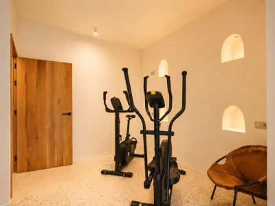 Dedizierter Fitnessbereich mit Crosstrainer und Heimtrainer auf Terrazzoboden, ergänzt durch beleuchtete Bogennischen und eine Holztür.