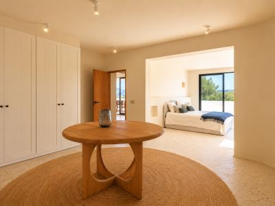 Dieser helle Übergangsbereich verfügt über einen runden Holztisch auf einem Juteteppich, flankiert von weißen Einbauschränken. Er führt zu einem Schlafzimmer mit grüner Aussicht.
