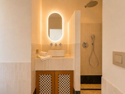 Dieses elegante Badezimmer bietet einen Aufsatzwaschtisch, einen ovalen, hinterleuchteten Spiegel und einen Holzschrank mit Gittertüren. Die Dusche hat einen Regenduschkopf.