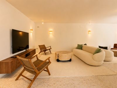 Ein moderner Wohnbereich mit geschwungenem cremefarbenem Sofa, rustikalem Holztisch und Korbsesseln auf Terrazzoboden, beleuchtet von warmen Wandleuchten.