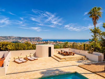 Großzügige Steinterrasse mit Pool, Sonnenliegen und erhöhtem Essbereich aus Holz, mit Panoramablick auf die Bucht von Jávea und die Küste unter blauem Himmel.