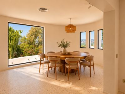 Heller Essbereich mit organischem Holztisch für acht Personen und Terrazzoboden. Große, geschwungene Fenster bieten Blick auf üppiges Grün und die ferne Landschaft.