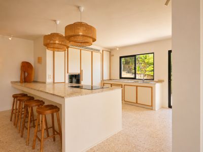 Moderne Kücheninsel mit integriertem Kochfeld, heller, gesprenkelter Arbeitsplatte und fünf Holzbarhockern. Geflochtene Pendelleuchten erhellen den Raum, mit Blick auf üppiges Grün im Freien.