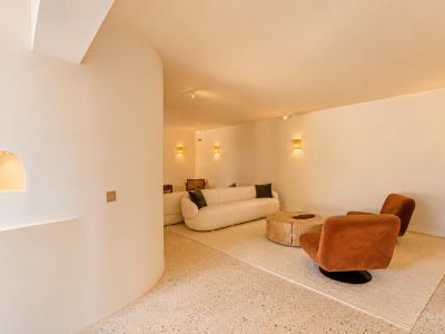 Moderner Loungebereich mit geschwungenem cremefarbenen Sofa, zwei rostfarbenen Sesseln und rustikalem Holztisch auf hellem Terrazzoboden. Geflochtene Wandleuchten sorgen für warmes Licht.
