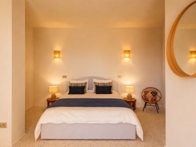 Schlafzimmer mit Doppelbett, Holznachttischen und Keramiklampen. Wandleuchten mit Raphia-Schirmen und ein runder Holzspiegel prägen den Raum. Terrazzoboden.