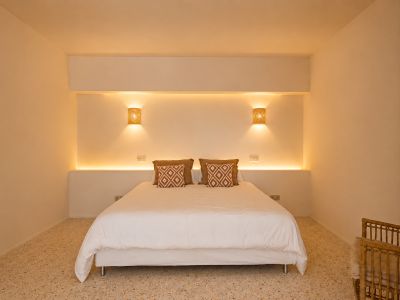 Schlafzimmer mit Doppelbett, integrierter LED-Kopfteilbeleuchtung und geflochtenen Wandleuchten. Terrazzoboden und gemusterte Kissen.