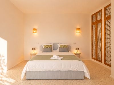 Schlafzimmer mit hellblau gepolstertem Bett, Terrazzoboden und warmer Beleuchtung durch geflochtene Wandleuchten. Rechts ein Holzschrank mit Gittertüren.