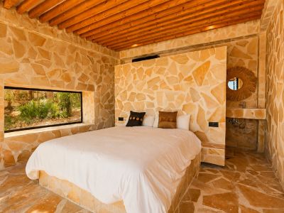 Schlafzimmer mit unregelmäßigen Natursteinwänden und -boden, Holzbalkendecke und einem horizontalen Fenster mit Gartenblick. Ein integriertes Steinwaschbecken mit Rattan-Spiegel ist sichtbar.