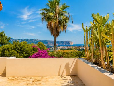Terrasse mit unregelmäßigem Steinboden und niedriger beiger Mauer. Bietet einen Kaktusgarten und Panoramablicke auf das Mittelmeer, die Küste und ein entferntes Kap bei blauem Himmel.