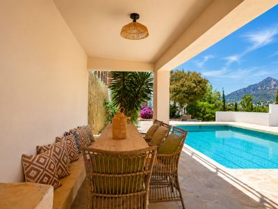 Überdachter Essbereich im Freien mit Holztisch und Korbstühlen, direkt am Pool gelegen. Panoramablick auf den Montgó und mediterrane Pflanzenwelt auf Steinboden.