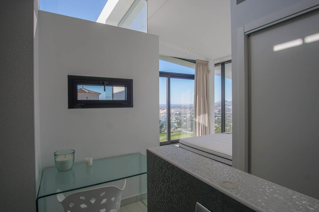 Arbeitsbereich mit Glasschreibtisch und horizontalem Fenster, der vom angrenzenden Schlafzimmer aus einen Panoramablick auf Meer und Stadt bietet.