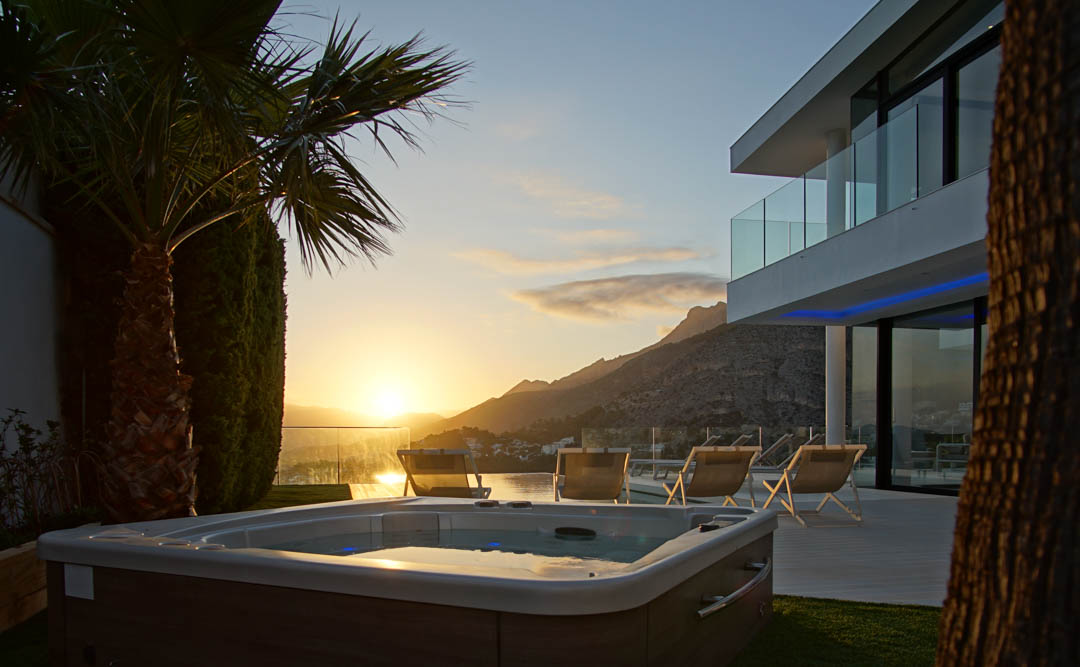 Außen-Jacuzzi mit Holzoptik auf Rasenfläche, vor einer modernen Villa mit Glasbalkonen. Panoramablick auf Berge und Meer bei Sonnenuntergang.