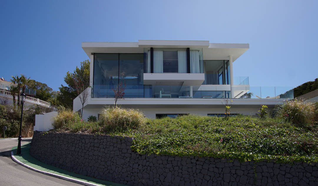 Außenansicht einer modernen dreistöckigen Villa mit weitläufigen Glasfassaden und Glasgeländern, erhöht über einer Steinmauer und Vegetation an einer geschwungenen Straße.