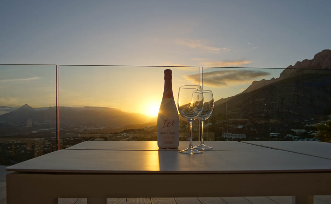 Außentisch auf einer Terrasse mit Glasgeländer, mit Rosé-Cava-Flasche und zwei Gläsern. Sonnenuntergang über Bergen und Tal, mit warmen Reflexionen des Abendlichts.
