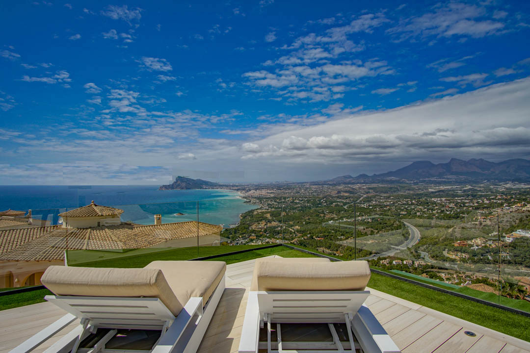Dachterrasse mit zwei weißen Sonnenliegen auf hellem Holzdeck und Kunstrasen, mit Panoramablick auf das Mittelmeer, die Küste von Altea und ferne Berge.