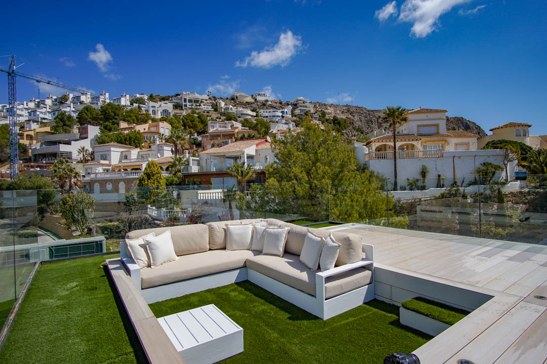 Dachterrassen-Lounge mit L-förmigem Sofa auf Kunstrasen, weißem Couchtisch und Glasgeländer. Bietet klare Aussicht auf den terrassierten Hang mit Wohnhäusern.