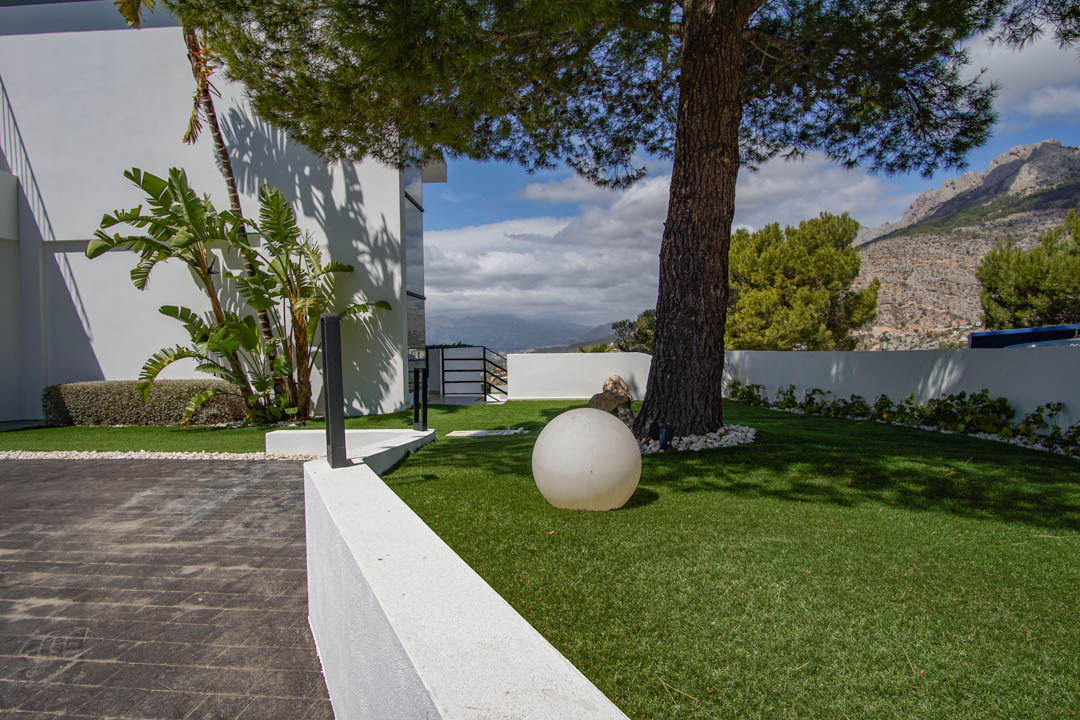 Der Außenbereich der Villa zeigt eine gepflasterte Auffahrt, Kunstrasen mit einer alten Kiefer, eine moderne Kugelleuchte und Bergblick.
