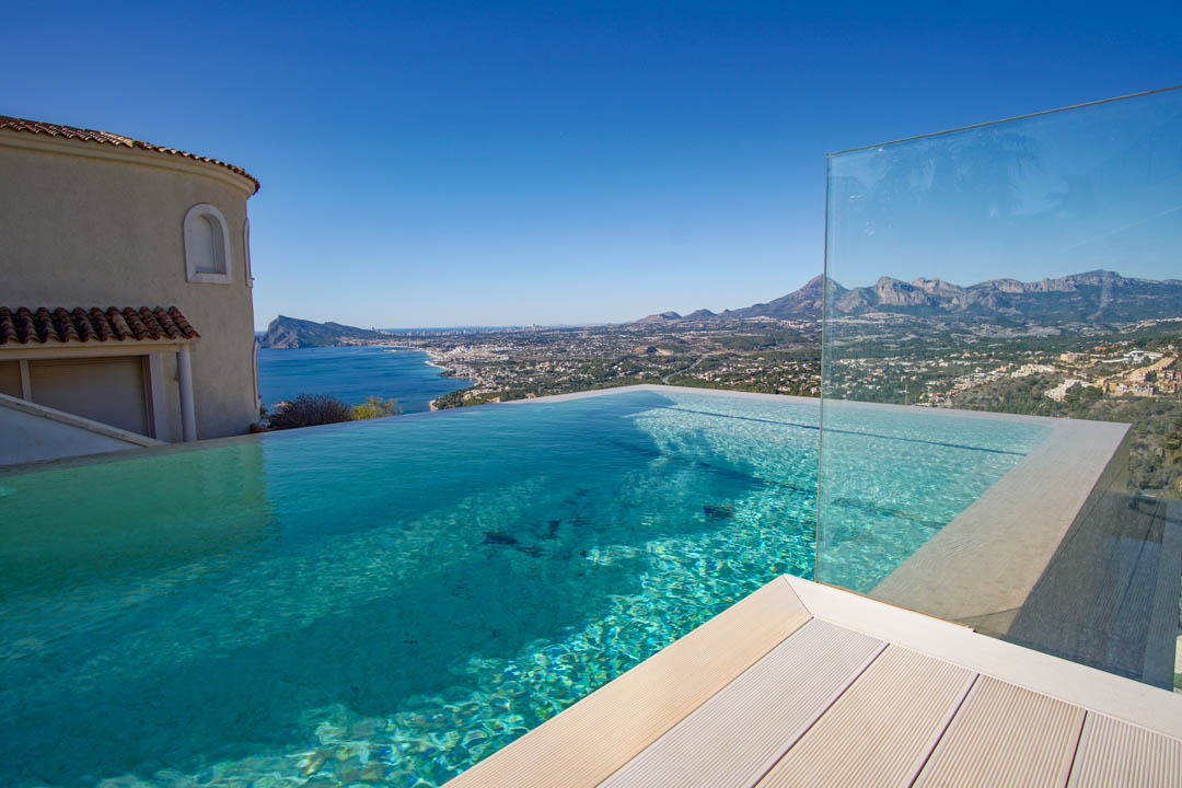 Der Infinity-Pool mit klarem Wasser, heller Decking und Glasbarriere bietet einen Panoramablick auf Meer, Küste und ferne Berge unter blauem Himmel.